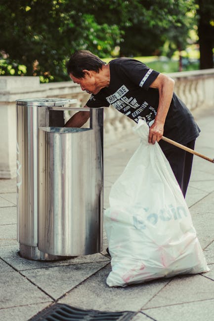 A middle-aged man with dark, slightly wavy hair is seen using a metal litter bin on an outdoor paved walkway, which features a curved refuse opening and a shiny, reflective surface. He is dressed in a dark T-shirt with white and blue text and graphics, and his posture indicates he is actively disposing of waste. In his left hand, he holds a large white plastic bag filled with rubbish, secured at the top, while his right hand is inserted into the bin, possibly to deposit waste. The scene is set in a well-maintained urban environment with a low stone or concrete balustrade on the background's right side and lush green foliage and trees visible behind, suggesting a park or a landscaped public space. The lighting appears natural, typical of daylight, emphasizing the clarity of the materials and textures, including the metallic sheen of the bin, the fabric of his clothing, and the texture of the plastic waste bag. This scene exemplifies a cleanup activity suitable for waste removal services such as those provided by Rubbish Removal Sutton, highlighting on-site rubbish collection and disposal methods distinct from council-provided waste management.