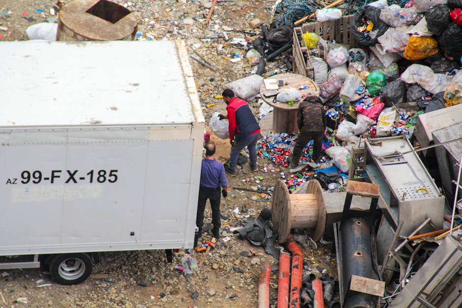 A small, blue, flatbed truck parked on the side of a street with its cargo area filled with a large pile of mixed rubbish, including cardboard boxes, black plastic bags, and miscellaneous waste materials. The truck's body shows signs of wear, with a slightly faded and chipped paint finish, and the wheels are painted red. The cargo is stacked high, extending above the truck's sides, and is loosely secured with ropes and cords. Behind the truck, there is a background of green trees with autumn-colored leaves, and nearby a green metal fence is visible. The scene suggests an outdoor waste collection or disposal activity, reflecting private rubbish removal efforts rather than a municipal service, consistent with the offerings of Rubbish Removal Sutton. Natural daylight illuminates the scene, with clear shadows cast by the truck and surrounding objects, emphasizing the urban environment's casual waste management context.