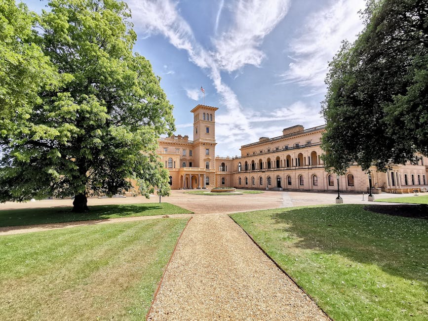 A broad pathway made of textured gravel leads through a well-maintained garden area toward a historic, light beige stone building with a prominent tower and arched windows. On either side of the pathway, there are lush green lawns bordered by mature trees. To the left, a large tree with vibrant green foliage extends its branches over the grass, while to the right, another tree provides shaded coverage. The building features a classical architectural style with decorative details and a roofline finished with a parapet, and a small flag is visible atop the tower. The sky above is partly cloudy with patches of blue and wispy clouds, creating an inviting outdoor environment. The scene appears to be in a landscaped public or private estate, with a focus on maintaining careful grounds management, consistent with on-site clearance or garden waste removal by a service such as Rubbish Removal Sutton, supporting independent disposal or landscape clearing efforts in the context of garden maintenance or estate management.